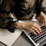 Kobieta z czerwonymi paznokciami pisze na laptopie obok notesu i długopisu
