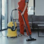 Pracownik w czerwonym kombinezonie odkurza salon żółtym odkurzaczem